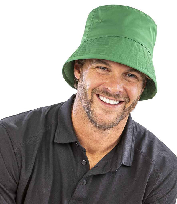 A man with a beard and mustache smiles at the camera, wearing a Result Recycled Result Core Recycled Bound Edge Bucket Hat in green, featuring an 8-stitch line brim and stitched eyelets, paired with a black collared shirt against a white background.