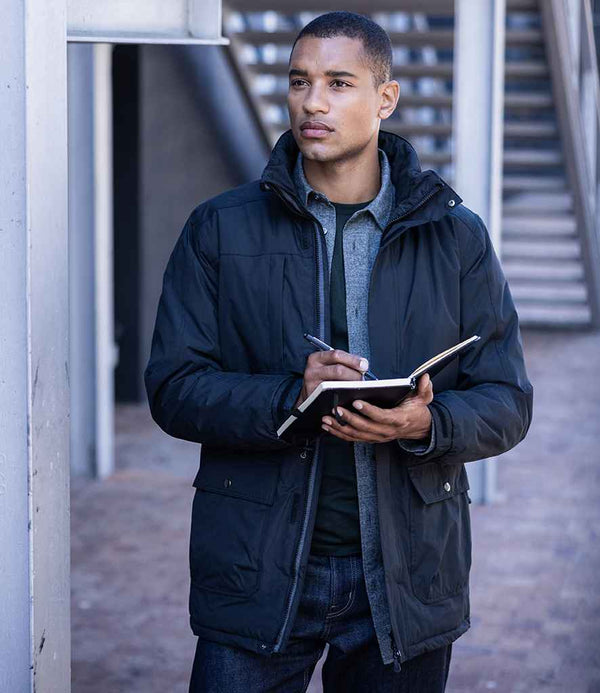 A person in a Regatta Professional Darby III Waterproof Insulated Parka Jacket made from recycled polyester stands outdoors, holding a notebook and pen, gazing sideways. Stairs and metal railings are visible in the background.