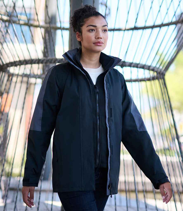 A person with curly hair wears a Regatta Professional Regatta Ladies Defender III 3-in-1 Jacket over a white shirt, standing outdoors before a metal wire structure, with blurred buildings and trees in the background.