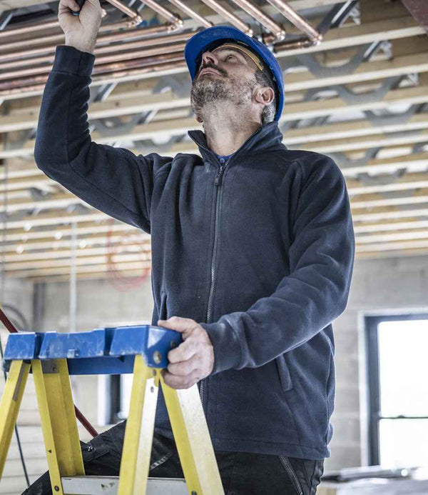 A construction worker wearing a Regatta Professional Sigma Heavyweight Fleece Jacket and a blue hard hat stands on a yellow ladder, inspecting overhead copper pipes in a building under construction.