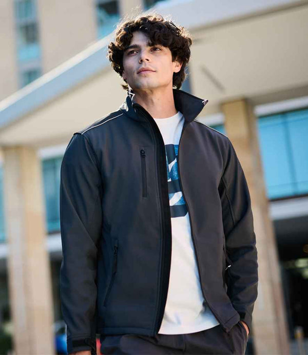 A young man with curly hair wears the Regatta Sandstorm Soft Shell Workwear Jacket by Regatta Professional over a graphic white T-shirt, standing outdoors in front of a modern building with tall windows and pillars, gazing slightly upward.