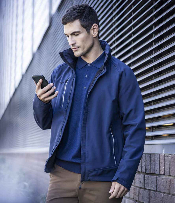 A man wearing a Regatta Professional Regatta Apex Soft Shell Jacket and brown pants stands outdoors by a metal-sided building, looking at his smartphone.