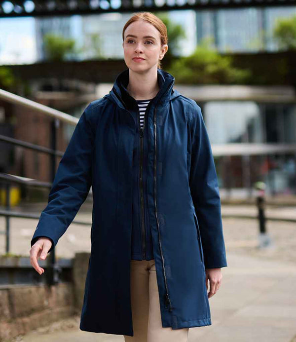 A woman stands outdoors near a railing, wearing the Regatta Professional Regatta Ladies Longline Kingsley 3-in-1 Jacket in navy blue over a striped shirt, with buildings and greenery blurred in the background.