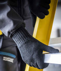 A person wearing Regatta Knitted Gloves by Regatta Professional grips a yellow ladder with one hand and a silver horizontal bar with the other; part of their dark jacket sleeve and pocket are visible.