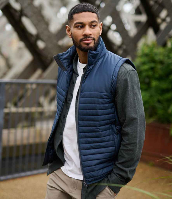 A Black man wears the Regatta Honestly Made Recycled Insulated Bodywarmer, a navy blue puffer vest, over a grey long-sleeved shirt and white T-shirt, paired with khaki pants.
