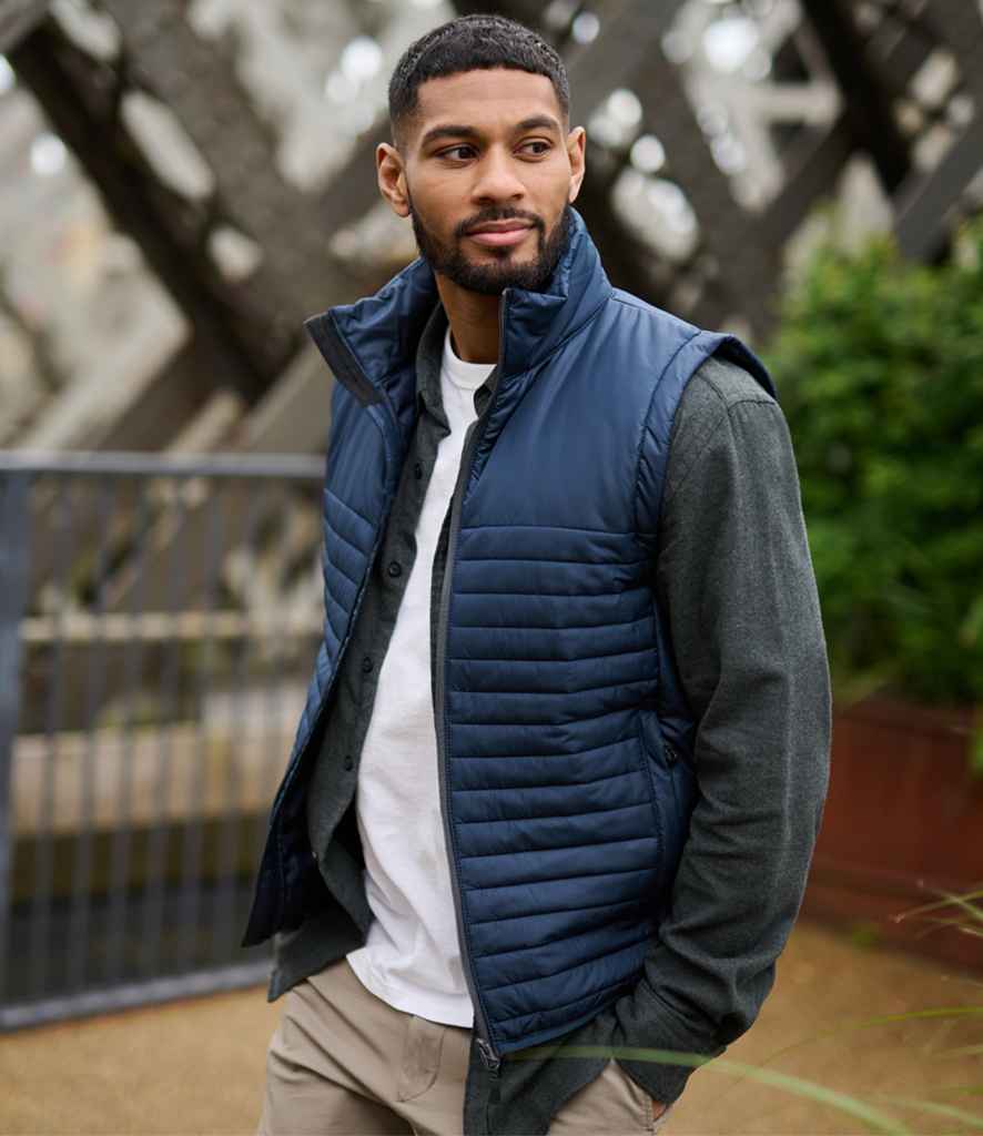 A Black man wears the Regatta Honestly Made Recycled Insulated Bodywarmer, a navy blue puffer vest, over a grey long-sleeved shirt and white T-shirt, paired with khaki pants.