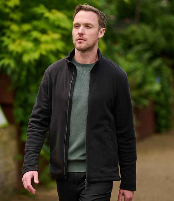 A man with short hair walks outdoors on a path, wearing the Regatta Honestly Made Recycled Micro Fleece Jacket over a green sweater, with greenery in the background.