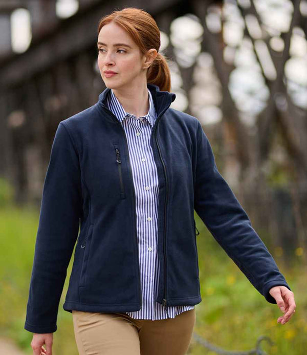 A person with long red hair in a ponytail is walking outdoors, wearing the Regatta Honestly Made Ladies Recycled Fleece Jacket over a blue-and-white striped shirt and khaki pants. Greenery and a wooden structure appear in the background.