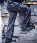 A person in Regatta Professionals Pro Stormbreak Waterproof Overtrousers with an elasticated waistband and boots steps onto rain-soaked concrete blocks, with visible water droplets on both the overtrousers and the blocks.