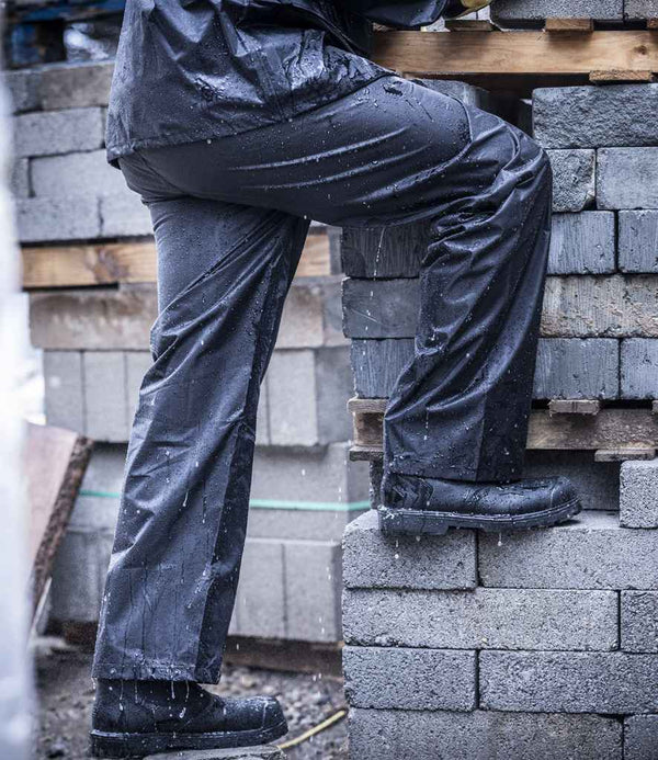 A person in Regatta Professionals Pro Stormbreak Waterproof Overtrousers with an elasticated waistband and boots steps onto rain-soaked concrete blocks, with visible water droplets on both the overtrousers and the blocks.