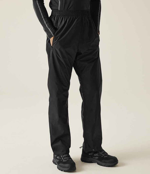 A person wears Regatta Pro Packaway Waterproof Breathable Overtrousers by Regatta Professional and black hiking shoes, standing against a plain light background.