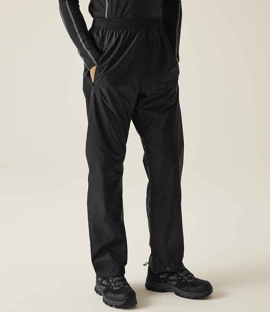 A person wears Regatta Pro Packaway Waterproof Breathable Overtrousers by Regatta Professional and black hiking shoes, standing against a plain light background.
