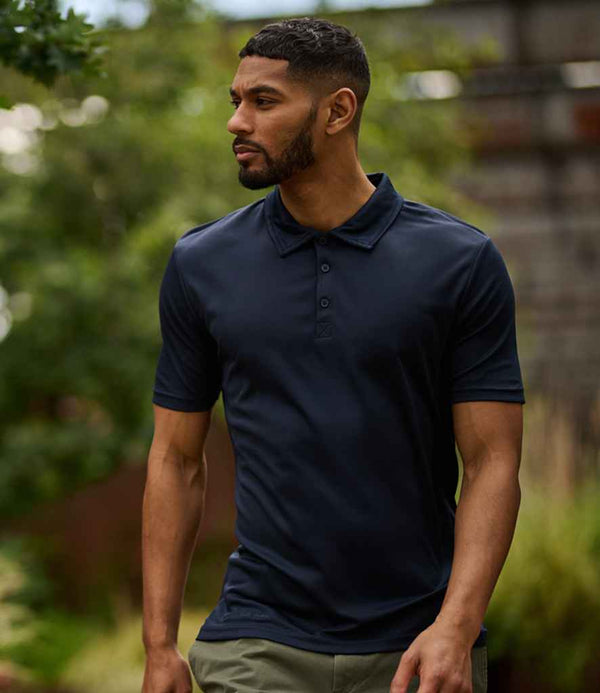 A man with short hair and a trimmed beard stands outdoors, wearing a Regatta Honestly Made Recycled Polo Shirt in navy blue with khaki pants. Trees and blurred greenery are visible in the background.