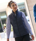 A person with curly hair walks outdoors, wearing a Regatta Professional Ladies Stage II Insulated Bodywarmer over a blue and white striped shirt and black pants. Blurred city buildings form the urban background.