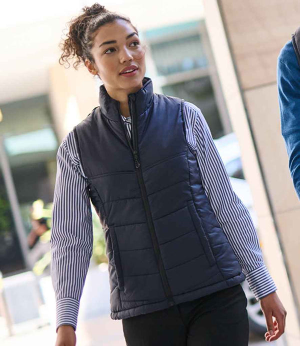 A person with curly hair walks outdoors, wearing a Regatta Professional Ladies Stage II Insulated Bodywarmer over a blue and white striped shirt and black pants. Blurred city buildings form the urban background.
