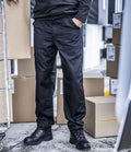 A person wearing Regatta Professional Regatta Action Trousers, a black long-sleeve shirt, and black work boots stands with one hand in their pocket, surrounded by cardboard boxes and a metal cart.