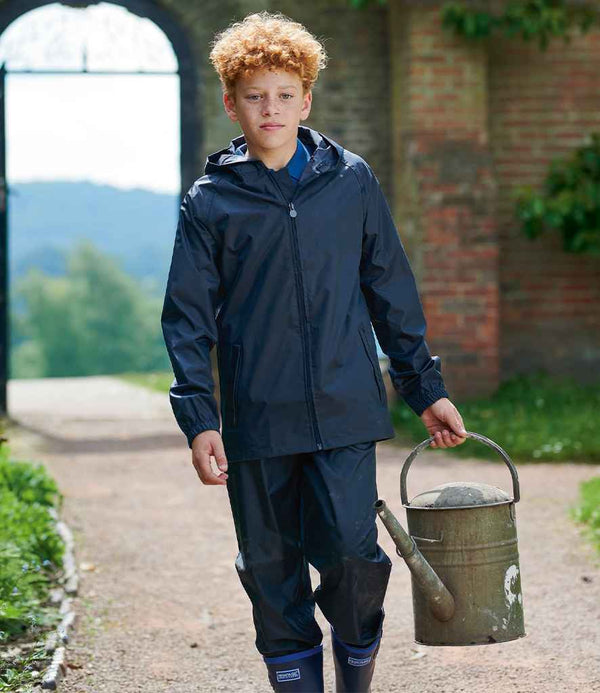A curly-haired boy wearing a Regatta Kids Pro Stormbreak Waterproof Jacket by Regatta Professional, matching pants, and rain boots walks on a path with a metal watering can. Greenery and a brick wall are in the background.