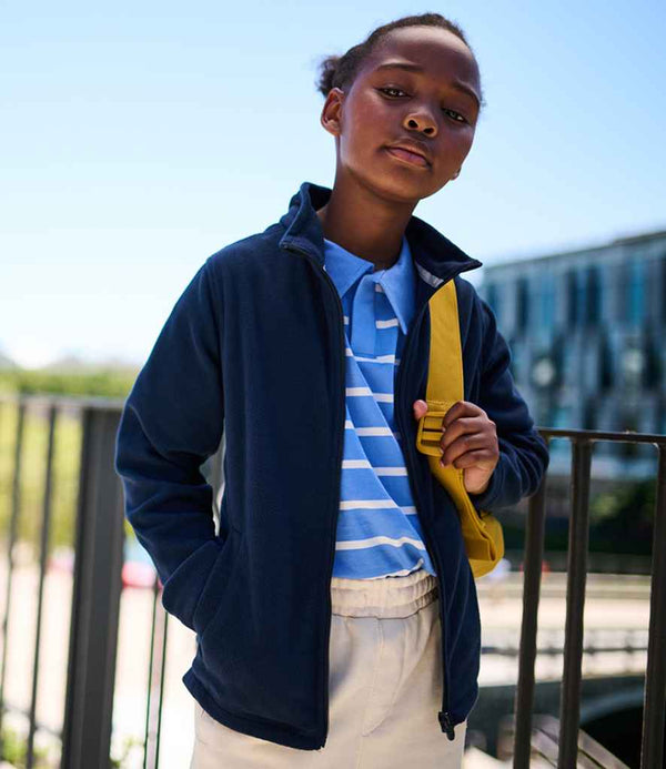 A young person stands outdoors with hands in their pockets, wearing the Regatta Kids Brigade II Micro Fleece Jacket by Regatta Professional, a striped polo, beige pants, and a yellow strap over one shoulder; a blurred building is in the background.