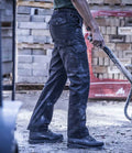 A person wearing Regatta Pro Cargo Trousers by Regatta Professional stands outdoors in sturdy black shoes, holding a handle. Stacked bricks or concrete blocks appear in the background, with focus on their lower body and legs.