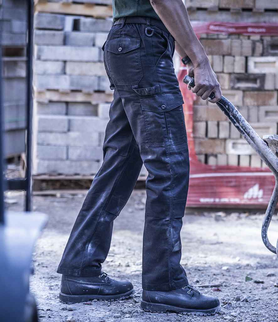 A person wearing Regatta Pro Cargo Trousers by Regatta Professional stands outdoors in sturdy black shoes, holding a handle. Stacked bricks or concrete blocks appear in the background, with focus on their lower body and legs.