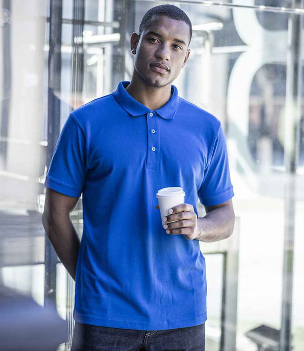 A man stands indoors by glass walls, wearing the Regatta Professional Regatta Classic Piqué Polo Shirt with a ribbed collar and three-button placket, paired with dark pants, and holding a white disposable coffee cup in his right hand.