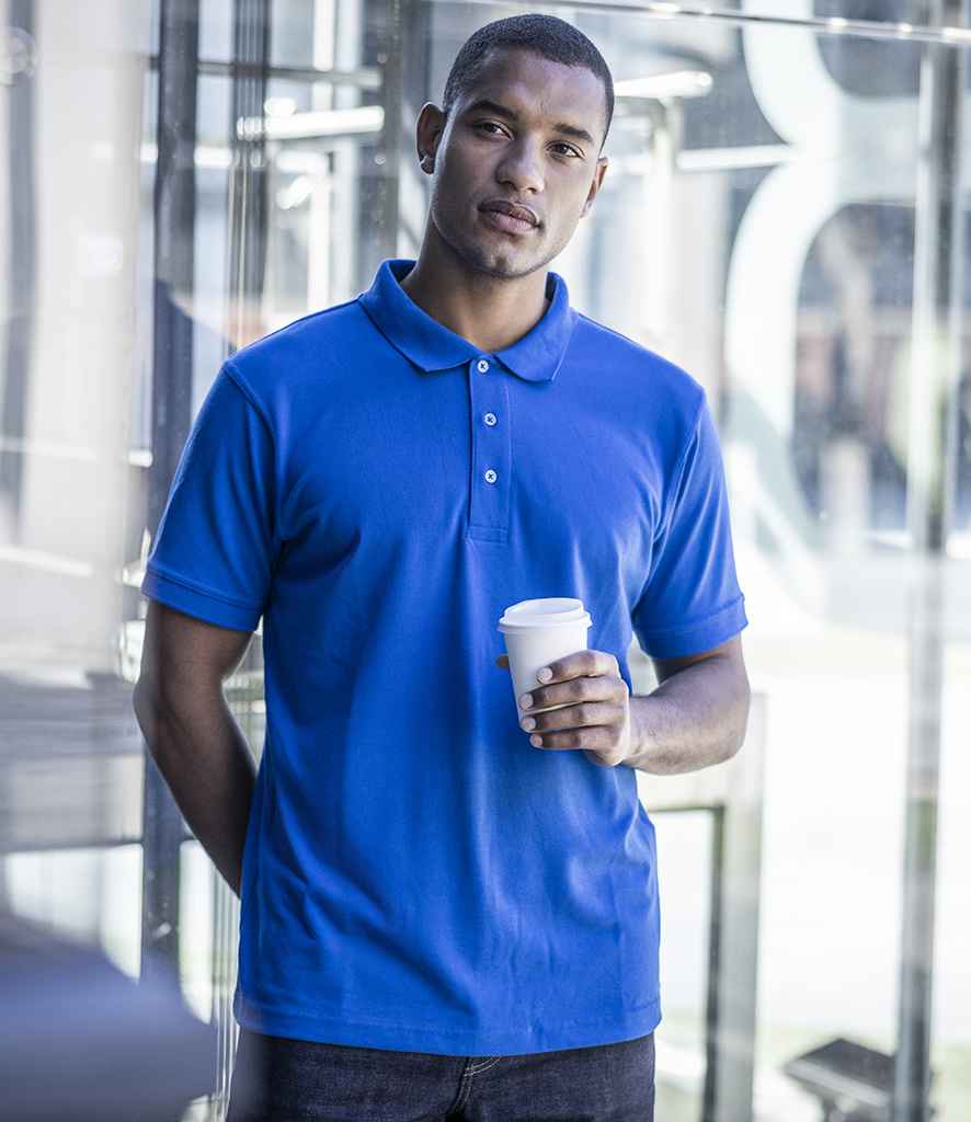 A man stands indoors by glass walls, wearing the Regatta Professional Regatta Classic Piqué Polo Shirt with a ribbed collar and three-button placket, paired with dark pants, and holding a white disposable coffee cup in his right hand.