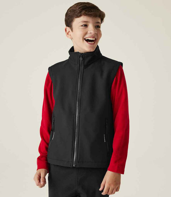 A boy with short brown hair smiles while wearing a Regatta Professional Kids Ablaze Soft Shell Bodywarmer over a red long-sleeve shirt and black pants, standing against a plain light-colored background.