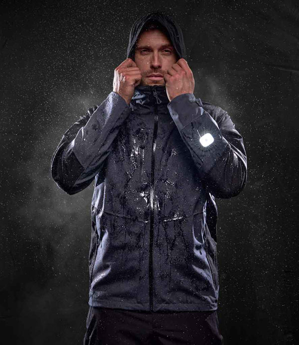 A person adjusts the hood of the Regatta X-Pro Beacon Brite Light Waterproof Shell Jacket by Regatta Professional, standing against a dark background with water droplets. A reflective sleeve detail enhances visibility in low light.