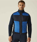 A man faces forward wearing the Regatta Professional Regatta E-Volve Unisex Knit Effect Stretch Bodywarmer with blue and black color blocks, high collar, zippered chest pockets, against a plain light-colored background.