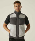 A man faces forward with his hands in his pockets, wearing a short-sleeved black shirt and the Regatta E-Volve Unisex Two Layer Soft Shell Bodywarmer by Regatta Professional. The background is plain and light-colored.