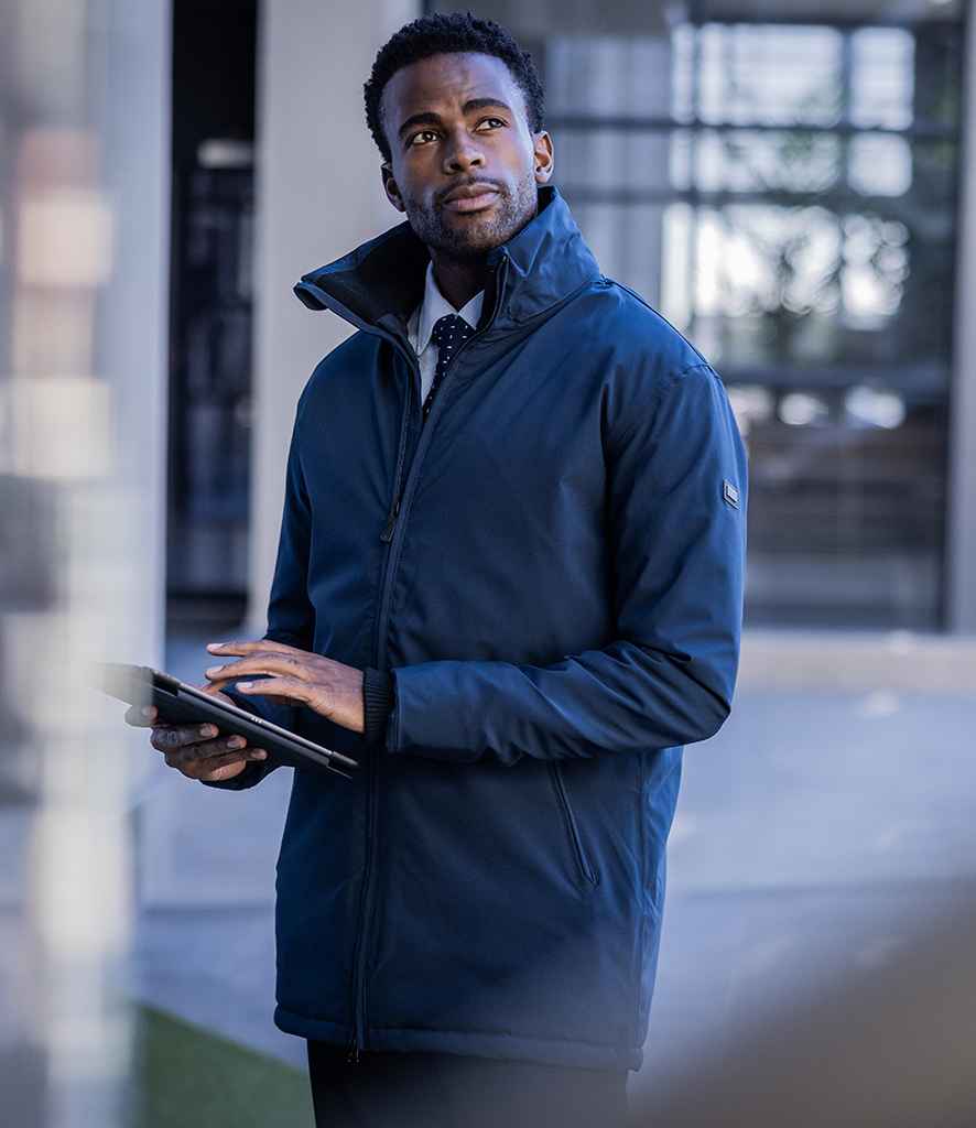 A man wearing the Regatta Professional Regatta Hampton Executive Jacket with Warmloft insulation stands outdoors holding a tablet, looking upward. The blurred background features glass and concrete structures.
