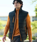 A man walks outside wearing the Regatta Professional Navigate Thermal Bodywarmer, a black showerproof vest with orange zippers, over an orange long-sleeve shirt. Greenery and a blurred building appear in the background.