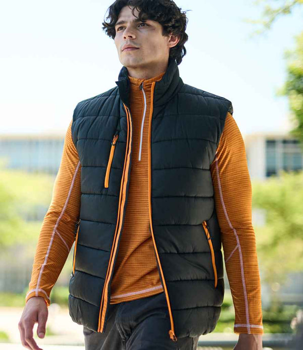 A man walks outside wearing the Regatta Professional Navigate Thermal Bodywarmer, a black showerproof vest with orange zippers, over an orange long-sleeve shirt. Greenery and a blurred building appear in the background.