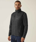 A man stands slightly turned, wearing the Regatta Navigate Hybrid Bodywarmer by Regatta Professional over a dark long-sleeve shirt and blue jeans, set against a plain light background.
