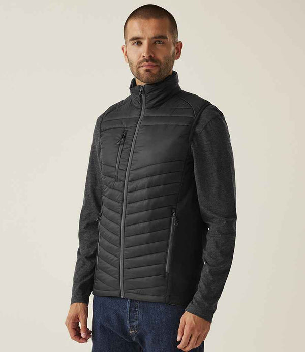 A man stands slightly turned, wearing the Regatta Navigate Hybrid Bodywarmer by Regatta Professional over a dark long-sleeve shirt and blue jeans, set against a plain light background.