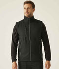 A man faces forward wearing the Regatta Navigate Two Layer Soft Shell Bodywarmer by Regatta Professional—a black, showerproof, recycled polyester vest with zippered pockets and a high collar—over a long-sleeve black shirt and pants. The background is plain and light.