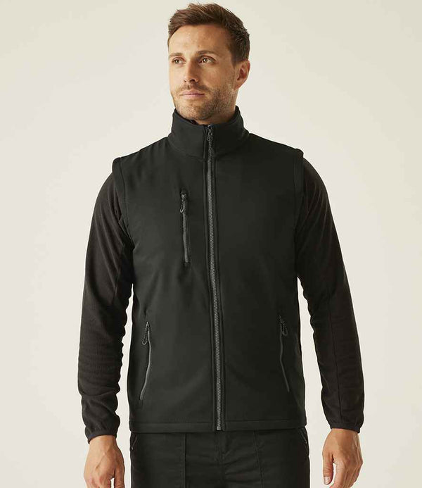 A man faces forward wearing the Regatta Navigate Two Layer Soft Shell Bodywarmer by Regatta Professional—a black, showerproof, recycled polyester vest with zippered pockets and a high collar—over a long-sleeve black shirt and pants. The background is plain and light.