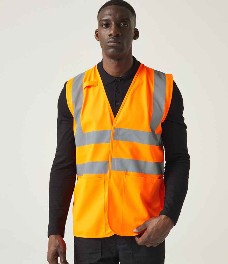 A person faces forward wearing the Regatta High Visibility Pro Supervisor Vest by Regatta High Visibility—an orange reflective safety vest with silver stripes that meets EN ISO 20471 standards—over a black shirt, against a plain light background.