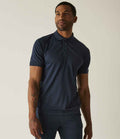 A man with a tattoo on his left forearm stands against a light background wearing the Regatta Professional Regatta Coolweave Piqué Polo Shirt in dark blue. The short-sleeve polo is made from quick-dry wicking performance fabric.