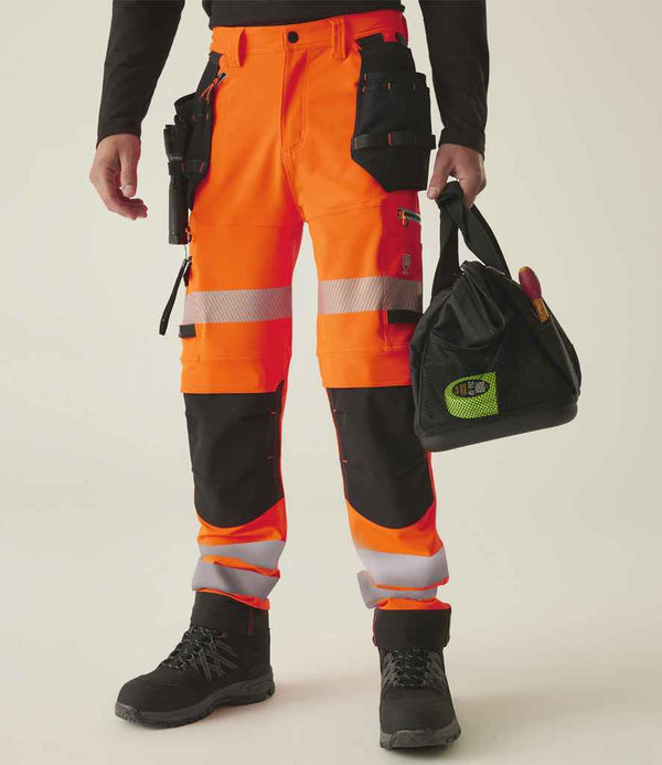 Person wearing Regatta High Visibility Infiltrate Stretch Holster Trousers in bright orange with black and reflective details, paired with black boots and a black long-sleeve shirt, holding a black tool bag. Face not visible.