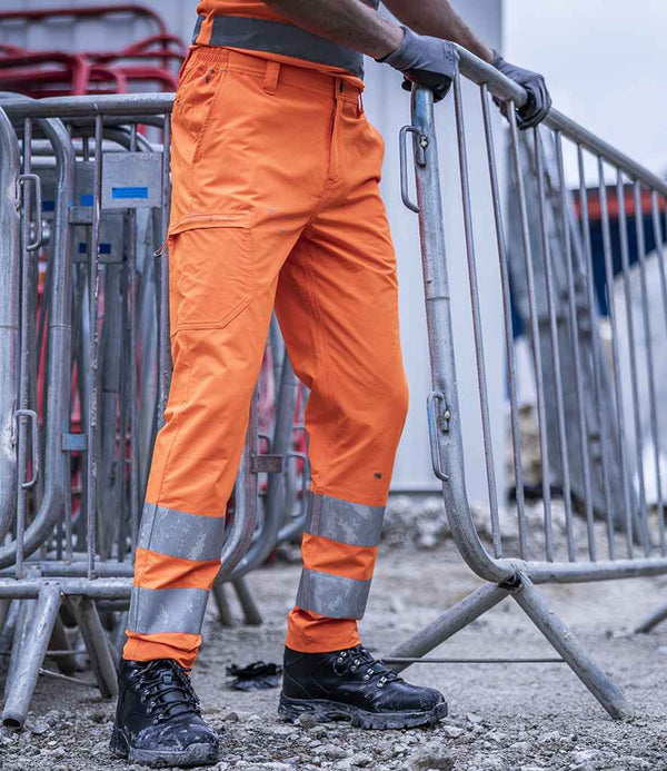 A person in Regatta High Visibility Pro Contract Stretch Action Trousers stands beside a metal barricade on a gravel construction site, wearing black work boots and gloves.