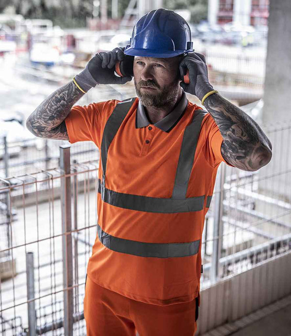 A construction worker in a Regatta High Visibility Pro Contract Polo Shirt and blue hard hat adjusts earmuffs at a construction site with safety fencing and equipment, meeting EN ISO 20471 standards.