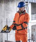 A construction worker in a Regatta High Visibility Pro Contract Ablaze Soft Shell Jacket, compliant with EN ISO 20471, and a blue hard hat holds a yellow nail gun and battery in an industrial setting with concrete walls and metal railings.