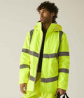 A person wearing the Regatta High Visibility Pro Contract Dover Parka by Regatta High Visibility, a neon yellow waterproof jacket with reflective gray stripes, stands against a beige background holding a black strap in their left hand.