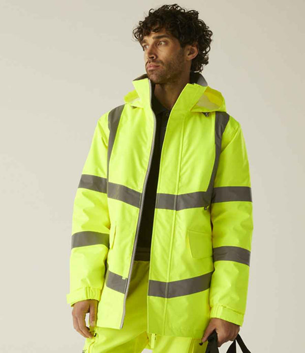 A person wearing the Regatta High Visibility Pro Contract Dover Parka by Regatta High Visibility, a neon yellow waterproof jacket with reflective gray stripes, stands against a beige background holding a black strap in their left hand.