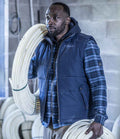 A man wears a Regatta Altoona Padded Bodywarmer by Regatta Professional and a blue plaid shirt, holding coils of white tubing in an industrial setting with additional coils visible in the background.