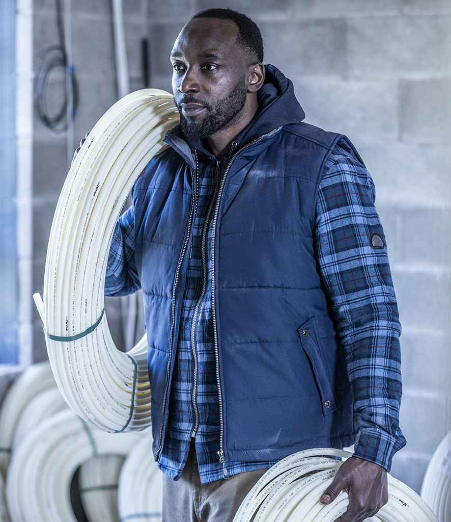 A man wears a Regatta Altoona Padded Bodywarmer by Regatta Professional and a blue plaid shirt, holding coils of white tubing in an industrial setting with additional coils visible in the background.