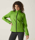 A person with curly hair wears the bright green Regatta Ladies Ablaze Printable Soft Shell Jacket by Regatta Professional and black pants, posing with one hand on their hip against a plain light background.