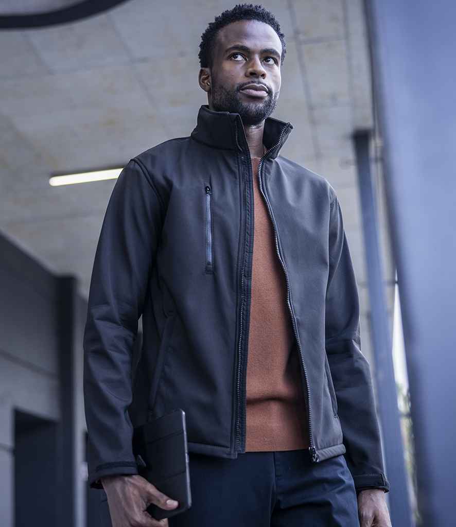 A man in a black Regatta Ablaze Three Layer Soft Shell Jacket by Regatta Professional stands outdoors with a black tablet, wearing a brown sweater. He has short hair and a beard, gazing upward in front of a modern building.