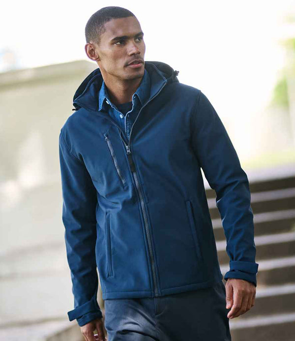 A man stands outdoors near steps, looking to the side, wearing the Regatta Professional Venturer Three Layer Hooded Soft Shell Jacket in blue with dark pants. The blurred background shows greenery and a light-colored wall.